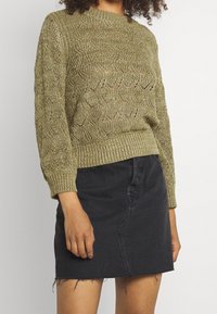 Suéter verde oliva de punto con patrón de tejido abierto, cuello redondo y puños acanalados, combinado con una minifalda de mezclilla negra con dobladillo deshilachado.
