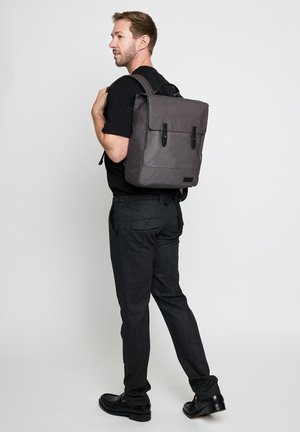 Man in black shirt and dark pants carrying a gray square backpack with two buckle straps, standing against a plain light background.