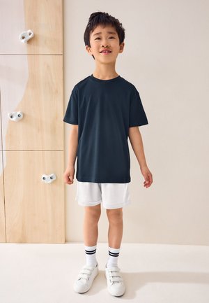 Jeune garçon portant un t-shirt noir, un short blanc et des baskets blanches, debout à l'intérieur contre un mur clair avec des prises d'escalade en bois.