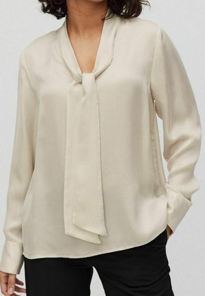 Mujer con blusa crema de manga larga con detalle de lazo en el cuello, combinada con pantalones negros, de pie con una mano en el bolsillo.