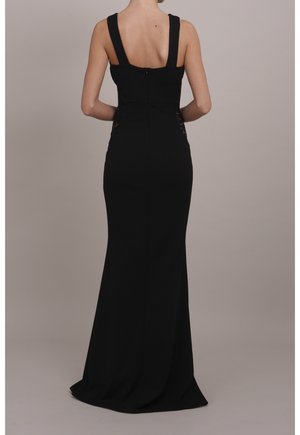 Femme debout, dos visible, portant une longue robe noire sans manches avec de larges bretelles et une taille cintrée.