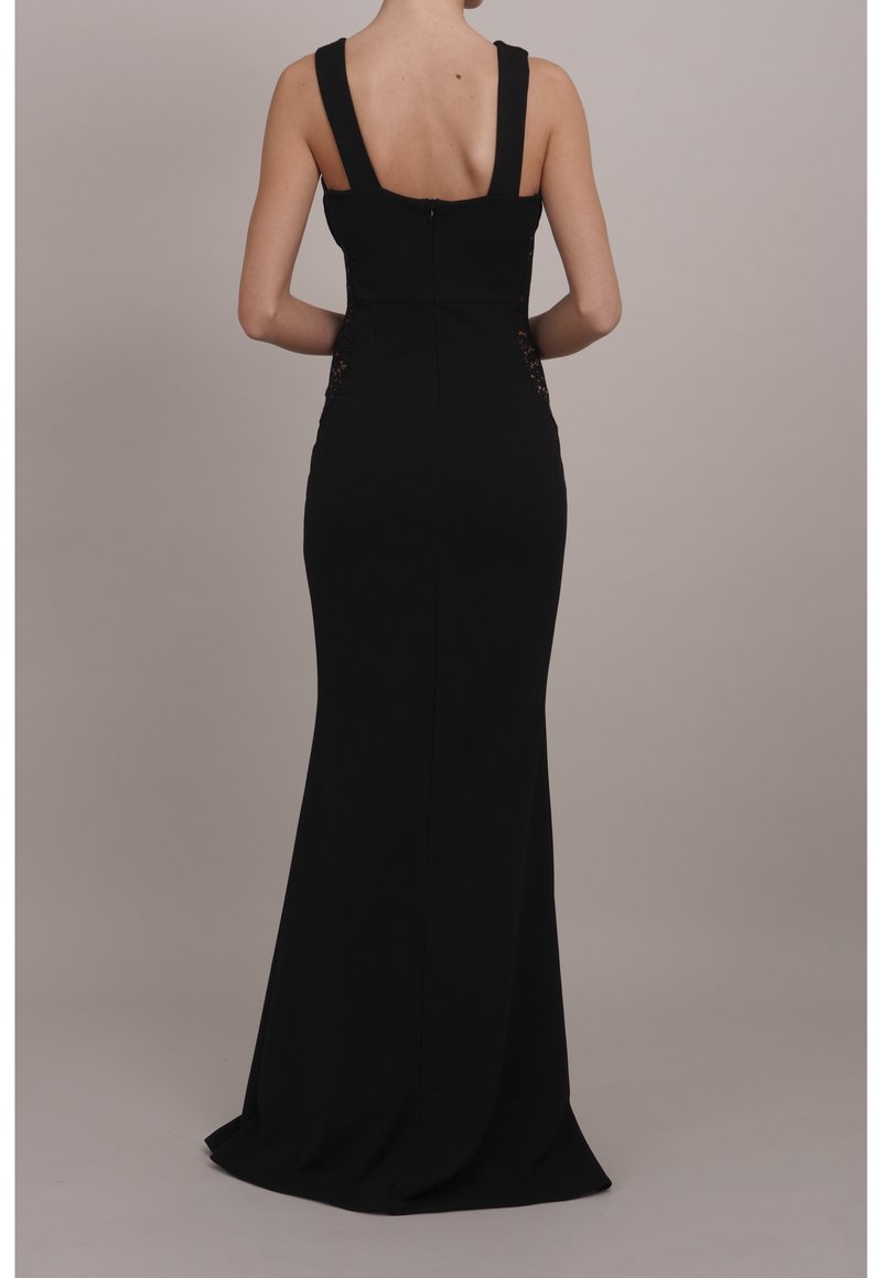 Femme debout, dos visible, portant une longue robe noire sans manches avec de larges bretelles et une taille cintrée.