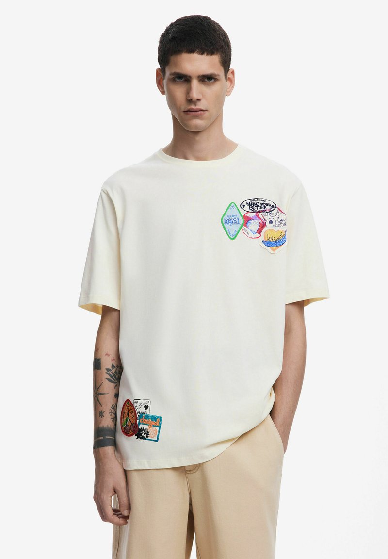 T-shirt corta a maniche corte color crema con vari toppe colorate che presentano disegni e testi sul petto e sull'orlo, abbinata a pantaloni beige.