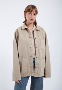 Beige oversized cotton jacket with a button front, two front pockets, and a relaxed fit, paired with light blue denim jeans.