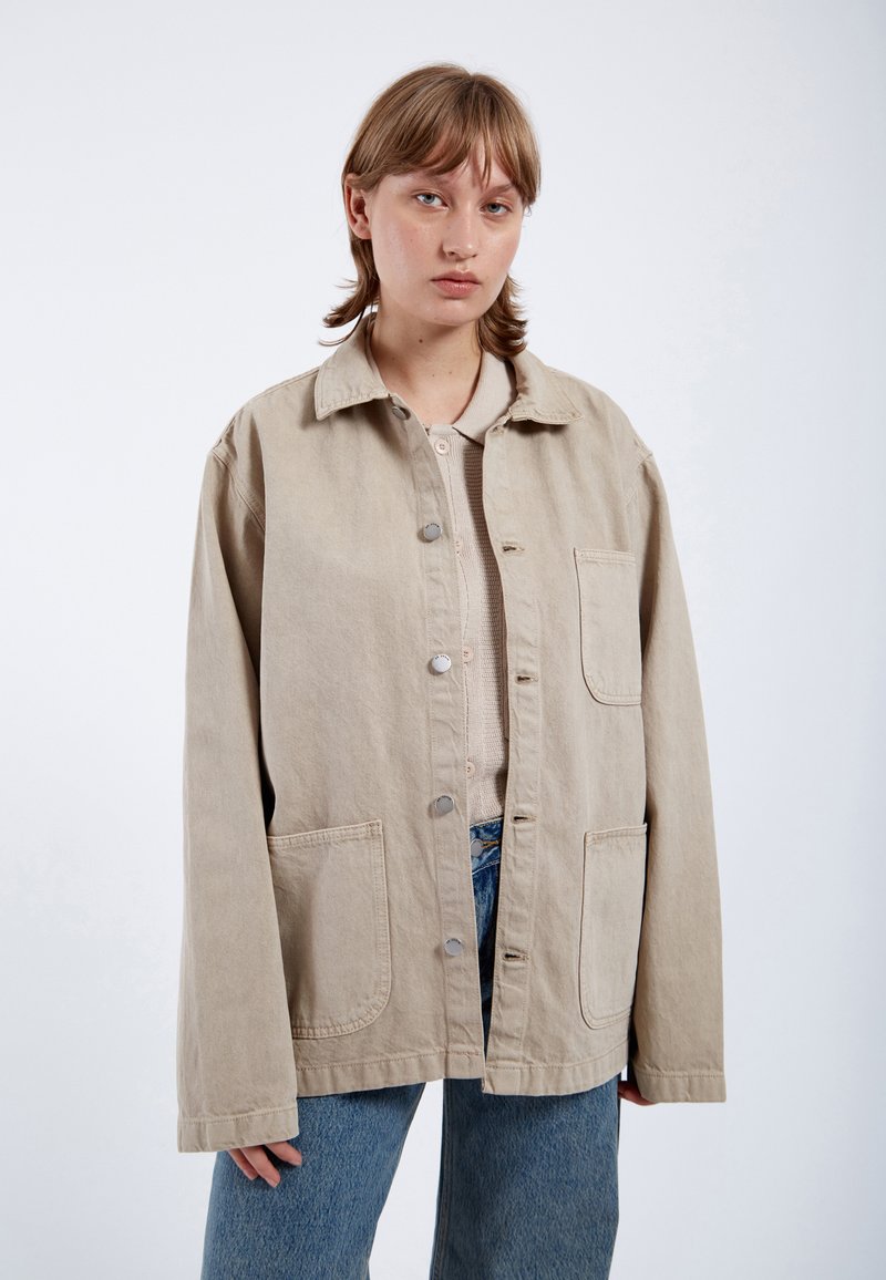 Beige oversized cotton jacket with a button front, two front pockets, and a relaxed fit, paired with light blue denim jeans.