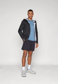 Vyras, dėvintis juodą su kapuce striukę, melsvą marškinius, juodus šortus, baltas kojines ir pilkas sportines batu, stovi prieš paprastą fono.