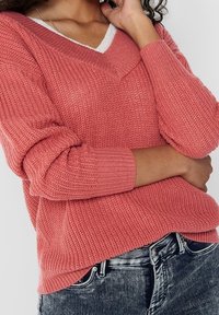 Koraalkleurige gebreide trui met een V-hals en lange mouwen, gekenmerkt door een textuurpatroon. Gedragen over een witte top en gecombineerd met donkere jeans.