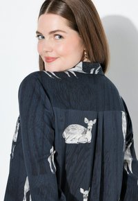 Donna con pelle chiara e lunghi capelli castani, che indossa una camicia blu scuro con illustrazioni di cervi bianchi, mentre guarda oltre la spalla e sorride leggermente.