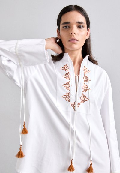 Tunique blanche brodée avec un col en V et des franges. Présente des broderies orange élaborées et de longues manches, fabriquée dans un tissu doux.