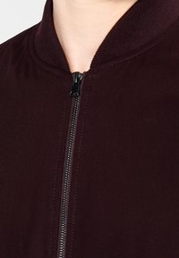 Close-up van een persoon in een donker kastanjebruine jas met een zwarte rits tot aan de nek, met een zichtbare geribde kraagdetails.