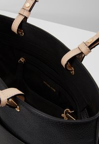 Mustav leather handbag, jossa on teksturoitu pinta, beige hihnat ja kultaiset yksityiskohdat. Sisäosassa on vetoketjullisia ja avoimia taskuja. Brändin logo näkyvissä.