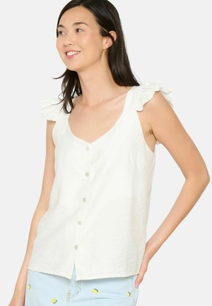 Blouse blanche sans manches avec des détails volantés aux épaules, boutonnage à l'avant et tissu texturé. Portée avec un short bleu clair avec des accents jaunes.
