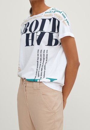 Osoba nosząca biały t-shirt z dużym czarnym i kolorowym napisem, połączonym z beżowymi spodniami, stojąca na jednolitym jasnym tle.