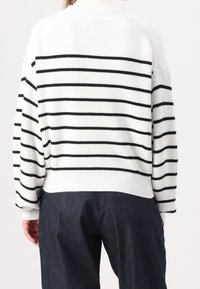 Biały sweter dziany w czarne poziome paski, o prążkowanej strukturze, luźnym kroju i wysokim kołnierzu. W zestawie z ciemnymi dżinsowymi spodniami.