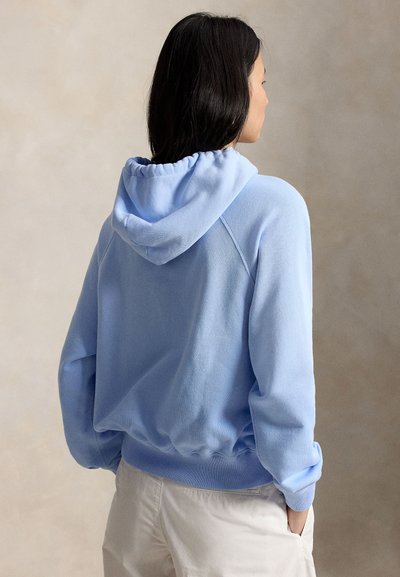 Femme aux longs cheveux noirs portant un sweat à capuche bleu clair et un pantalon de couleur claire, debout les mains dans les poches, de dos.