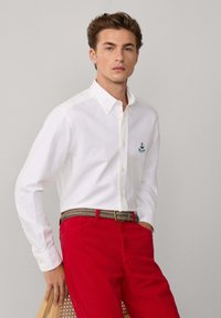 Joven vestido con una camisa blanca de botones con un pequeño emblema, pantalones rojos y un cinturón trenzado, de pie con una mano sobre una silla.