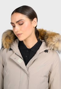 Beige zimní bunda s kapucí podšitou kožešinou, na zip, a pod ní černá košile na knoflíky. Obsahuje prošívání a nášivku s logem.