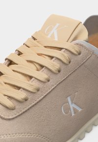 Μπεζ σουέτ αθλητικό παπούτσι με κορδόνια στον ίδιο τόνο, με λευκά λογότυπα "cK" στο πλάι και στη γλώσσα, και ελαφριά σόλα με ραβδώσεις.