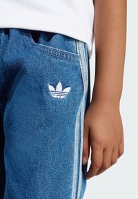 Pantaloni di jeans blu scuro con accenti a tre strisce bianche e un piccolo logo trifoglio ricamato sulla tasca anteriore.