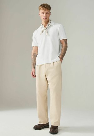 Junger Mann mit Tätowierungen, der ein weißes kurzärmeliges Hemd, beige Hosen, braune Schuhe und ein gemustertes Halstuch trägt und vor einem schlichten Hintergrund steht.