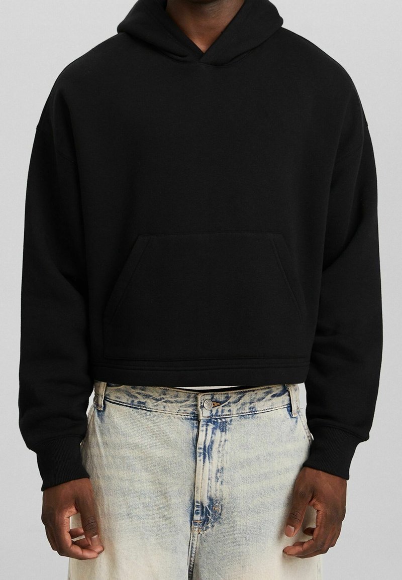 Sweat à capuche court noir avec poche avant porté par une personne en jean bleu clair délavé à l'acide, visible du cou aux hanches sur fond uni.