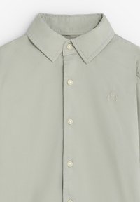 Camisa abotonada gris claro con cuello puntiagudo, que presenta un discreto logo bordado y pequeños botones en crema. Textura suave, diseño casual.