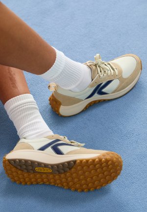 Baskets de sport beige et blanches avec des accents bleu marine, dessus en suède texturé et en cuir lisse, semelle en caoutchouc avec motifs de traction, associées à des chaussettes blanches.