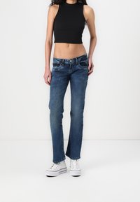 Top nero cropped senza maniche abbinato a jeans in denim blu con taglio dritto e tasche frontali; indossato con sneakers bianche.