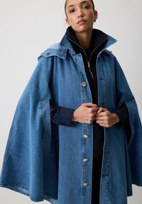 Denim kappe med højt krave, knaplukning foran og klaplommer. Lys blå farve med en let struktur og brede ærmer.