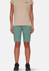 Lyse beige kortærmet t-shirt med trykt tekst, parret med knælange mintgrønne shorts. Sorte vandresko med tekstureret overflade.