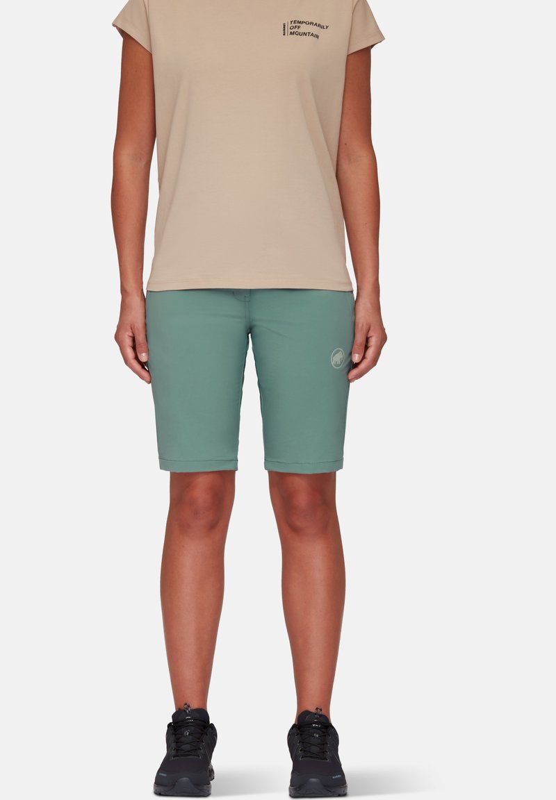 Lyse beige kortærmet t-shirt med trykt tekst, parret med knælange mintgrønne shorts. Sorte vandresko med tekstureret overflade.