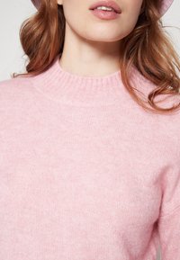 Femme à la peau claire et aux cheveux auburn ondulés porte un pull col côtelé rose pâle, dévoilant le cou et une partie des lèvres sur un fond uni.