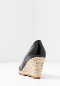 Zapato de tacón cuña de charol negro con una suela de yute trenzado, que presenta una punta afilada y un diseño curvado para un estilo adicional.