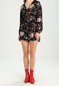 Robe noire à fleurs avec des manches longues, décolleté en V et accents en dentelle à la taille. Associée à des bottines rouges brillantes.