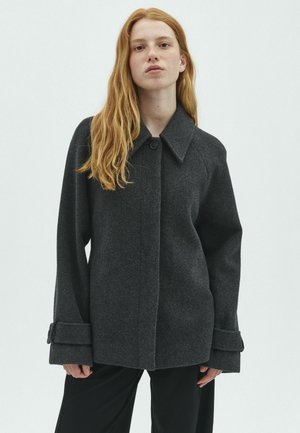 Jeune femme aux longs cheveux roux portant un manteau en laine gris foncé boutonné avec de larges poignets et un pantalon noir, debout devant un fond blanc.