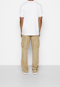 Mies seisoo selkä kameraan päin, päällään yksinkertainen valkoinen t-paita, vaaleat cargo-housut ja valkoiset lenkkarit vaaleaa taustaa vasten.