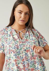 Blumenbluse mit kurzen Ärmeln, mit einem hellen Hintergrund, mehrfarbigen Blumen, einem Schlüsselloch-Ausschnitt und einer Schleifen-Detail am Kragen.