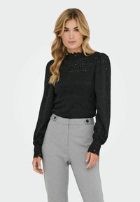 Juoda nėriniuota palaidinė su aukštu kaklu ir ilgomis rankovėmis. Turi tekstūruotą audinį ir banguotus rankogalius. Derinama su šviesiai pilkomis pritaikytomis kelnėmis.