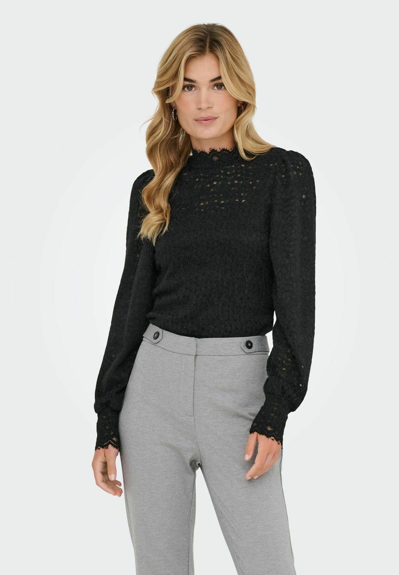 Juoda nėriniuota palaidinė su aukštu kaklu ir ilgomis rankovėmis. Turi tekstūruotą audinį ir banguotus rankogalius. Derinama su šviesiai pilkomis pritaikytomis kelnėmis.