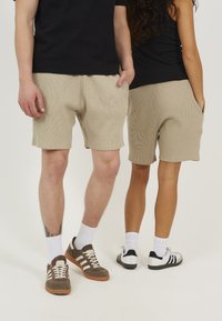 Des shorts beiges texturés avec des poches avant, associés à des sneakers marron et blanc et des sneakers noir et blanc. L'arrière-plan est entièrement blanc.