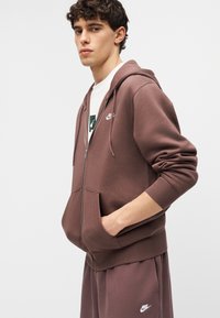 Jonge man die een bruine Nike hoodie met rits en bijpassende joggingbroek draagt, met één hand in de zak, kijkend naar de camera.