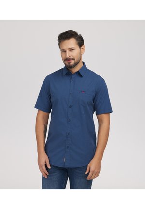 Mężczyzna z krótką brodą, ubrany w niebieską kraciastą koszulę na guziki z krótkim rękawem i ciemne dżinsy, stojący na tle jednolitego jasnego tła.