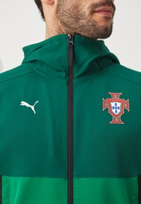 Zaļš jaka ar rāvējslēdzēju, izgatavota no gludas auduma, ar kapuci, Puma logotipu un Portugāles nacionālās komandas emblēmu ar sarkaniem un baltiem akcentiem.