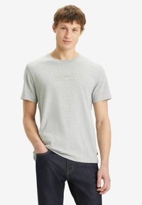 Camiseta de algodón gris con cuello redondo y mangas cortas, que presenta un logo de Levi's en relieve en la parte frontal. Combinada con jeans de mezclilla oscura.