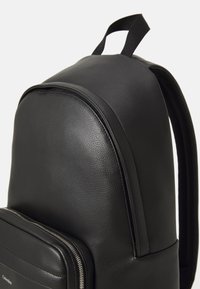 Sac à dos noir en cuir texturé, avec un sommet arrondi, une poche avant plate avec fermeture éclair et des bretelles doubles.