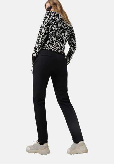 Schwarzer Pullover mit Muster und weißem Blumenmuster, schwarze Hose und beige Turnschuhe, Rückansicht. Trägt spiegelnde Sonnenbrille.