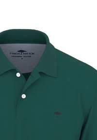 Zelená polo košeľa vyrobená z prémiovej bavlny, s golierom, tromi gombíkmi a malým hnedým logom stromu na hrudníku.