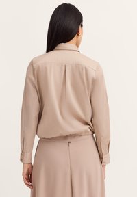Beige Bluse mit langen Ärmeln, Kragen, einem Rückenschlitz und taillierter Taille. Glatte Textur mit geknöpften Manschetten.
