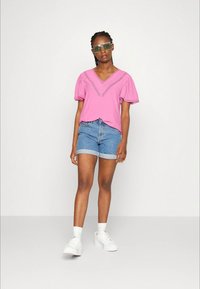 Top de algodão rosa com decote em V e mangas bufantes, combinado com calções de ganga azul claro e ténis brancos. Óculos de sol transparentes verdes.