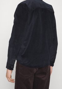 Granatowa koszula sztruksowa z tylną wstawką na plecach, długimi rękawami i mankietami na guziki, zestawiona z ciemnobrązowymi spodniami sztruksowymi.